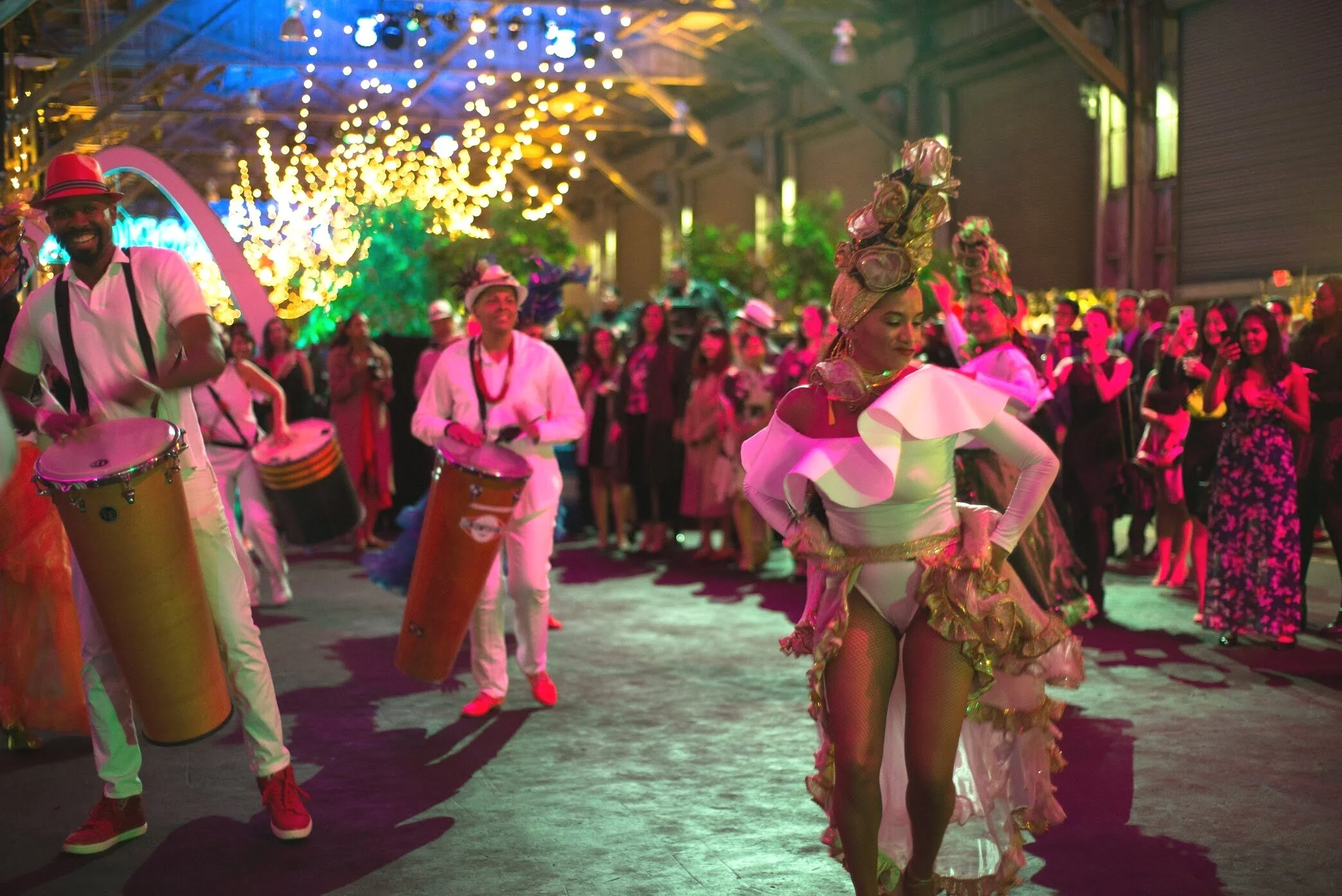 A lively indoor corporate event in San Francisco featuring a drum band and a female dancer in an extravagant floral headpiece. The drummers wear white and red as the colorful crowd enjoys vibrant entertainment under sparkling lights.