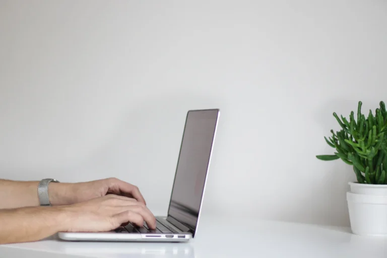 A person types on a silver laptop at a white desk. Only their hands and forearms are visible. To the right, a small green potted plant adds to the simple, minimalistic workspace—perfect for using the latest virtual meeting technology from your brand.