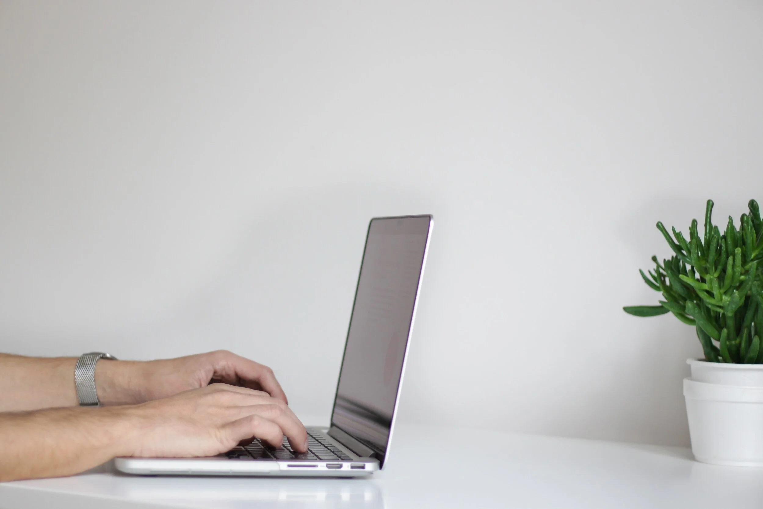 A person types on a silver laptop at a white desk. Only their hands and forearms are visible. To the right, a small green potted plant adds to the simple, minimalistic workspace—perfect for using the latest virtual meeting technology from your brand.