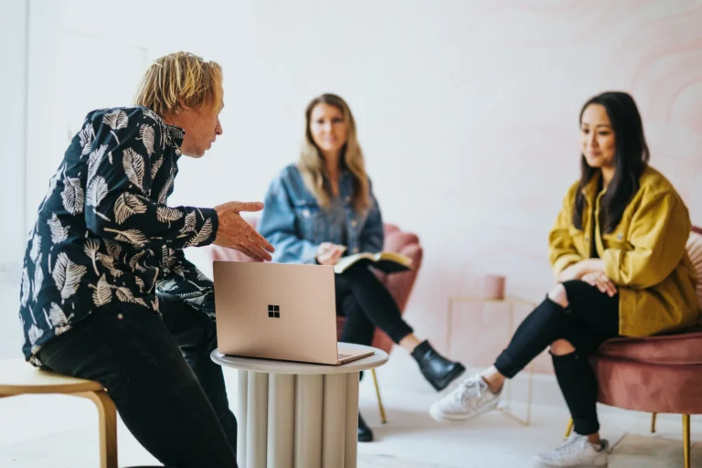 Three people sit indoors around a small table with a laptop, appearing engaged in a casual meeting or discussion about a virtual event. One gestures while talking, facing two others who listen attentively against a light pink background.