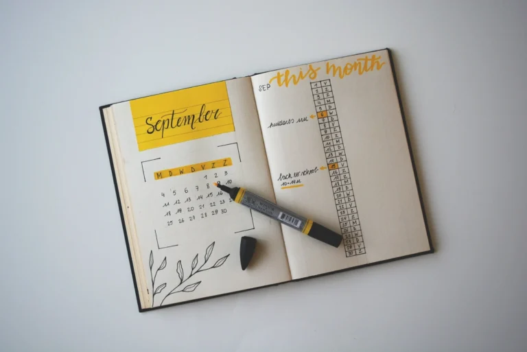 An open planner displays a September calendar on the left with yellow accents and a floral doodle. On the right, this month is written in yellow script above a habit tracker—perfect for tracking your virtual event planning time. A gray-and-yellow marker and black eraser rest on the pages.