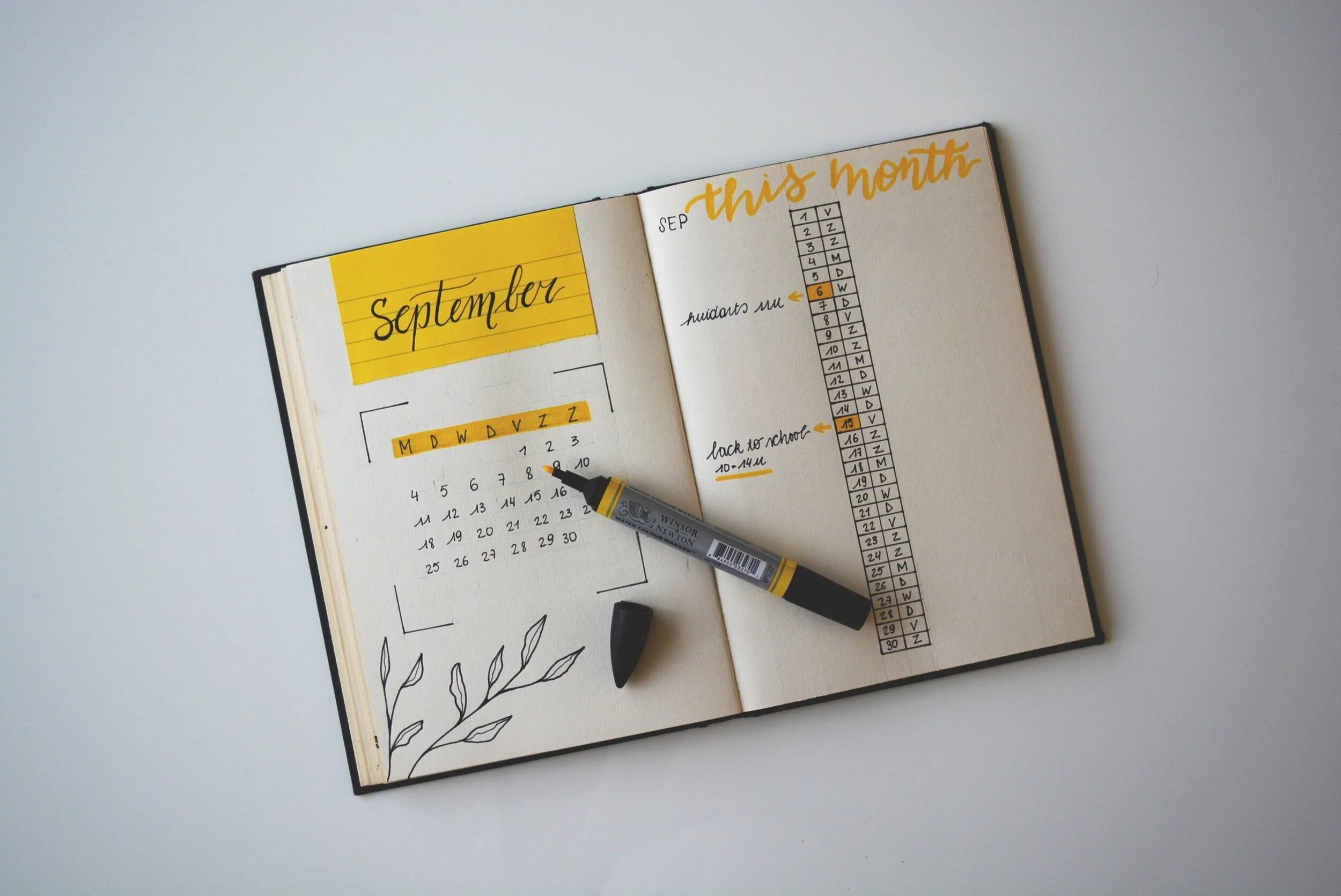 An open planner displays a September calendar on the left with yellow accents and a floral doodle. On the right, this month is written in yellow script above a habit tracker—perfect for tracking your virtual event planning time. A gray-and-yellow marker and black eraser rest on the pages.