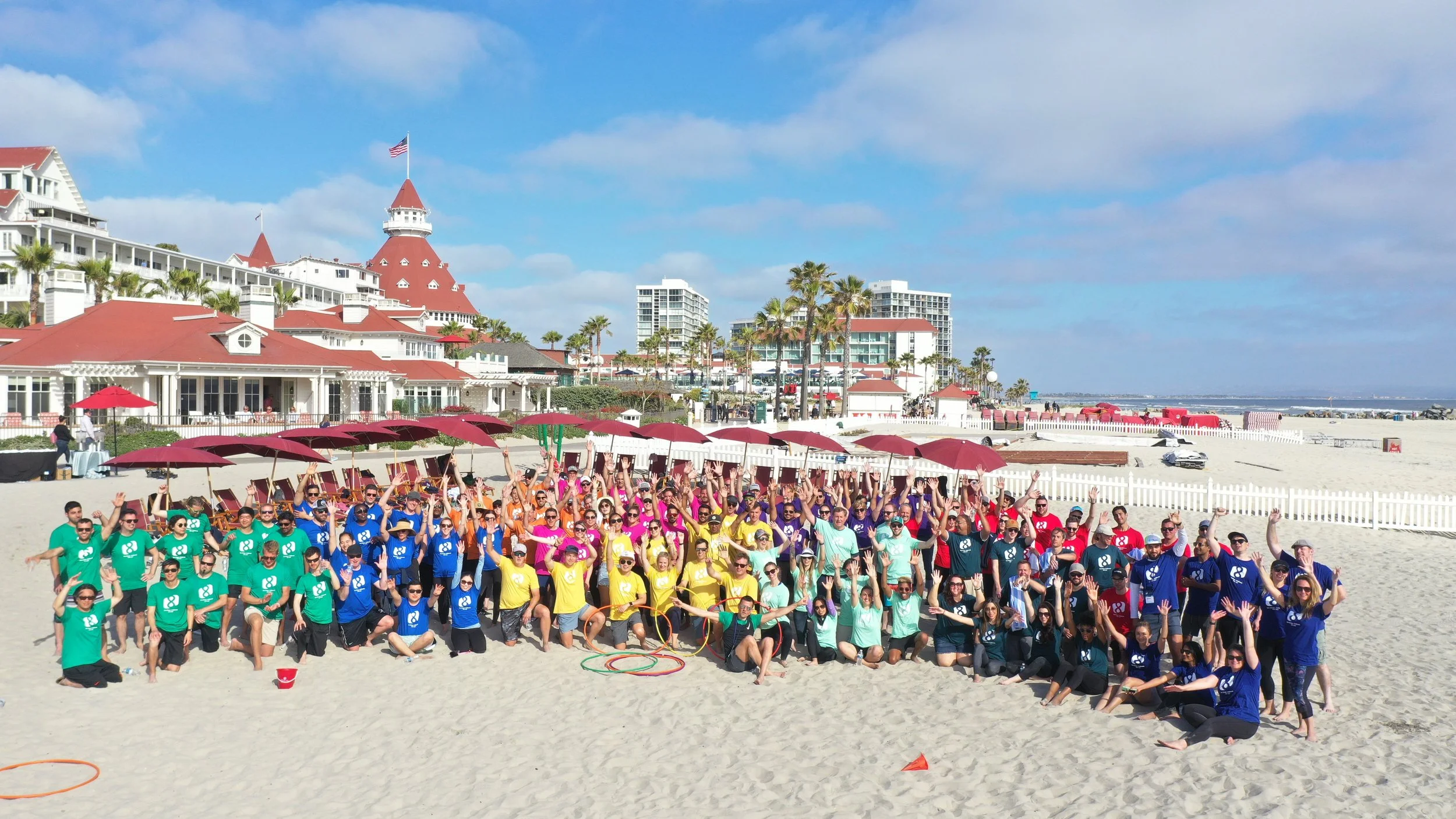 A large group, organized by a full-service event company, wearing color-coordinated team shirts, pose with raised arms on a sandy beach. Behind them are red-roofed buildings, palm trees, blue sky with clouds, red umbrellas, and a white fence.