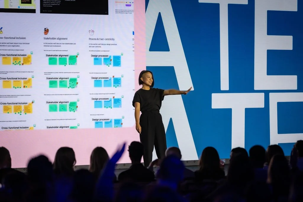 A person in black clothing stands on stage gesturing with one hand raised, addressing an audience—an example of effective event management. Behind them is a large, colorful presentation slide with diagrams and text, partially obscured by oversized blue and white lettering.