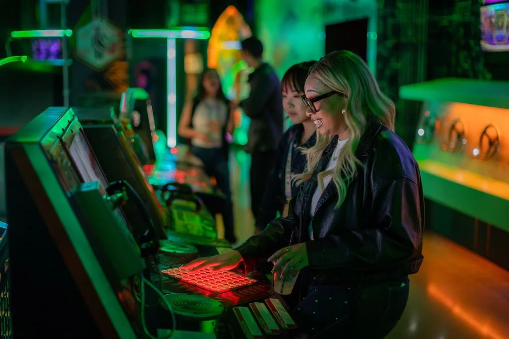 Two women in dark jackets smile and play arcade games in a neon-lit room. With event management creating a lively, futuristic atmosphere, glowing green and red lights illuminate the scene while more people enjoy colorful screens in the background.