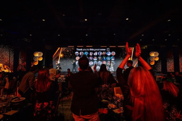 People in evening wear stand and clap at a dimly lit awards event. Showcasing expert event planning, a speaker stands near a large screen displaying team headshots with “Thank you to this great crew.” Tables with food and drinks fill the foreground.