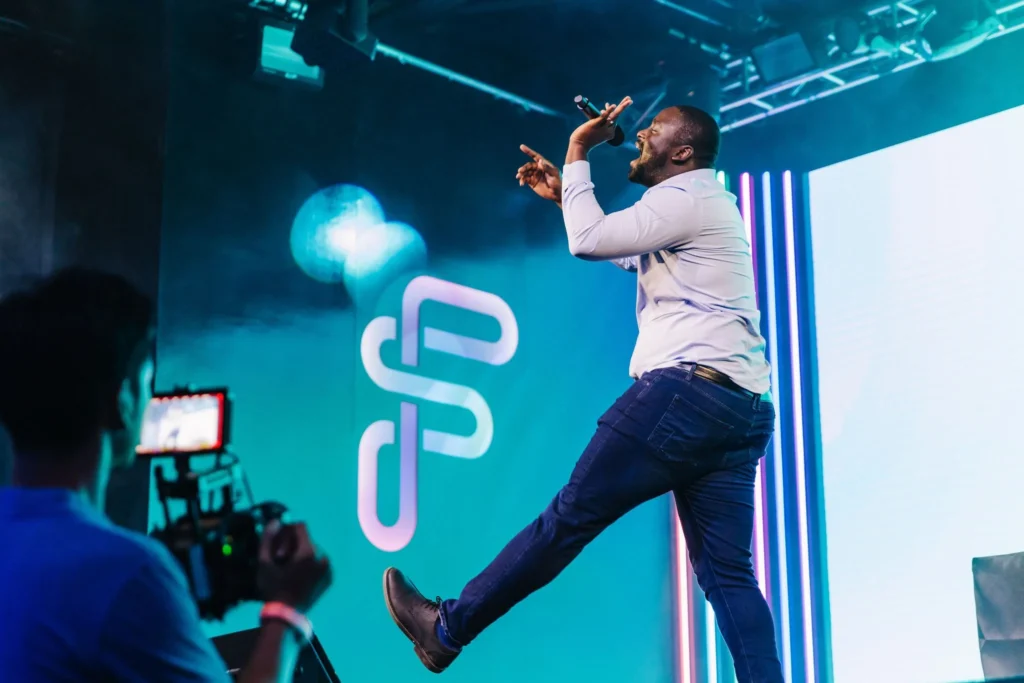 A man in a light shirt and jeans energetically performs on stage, holding a microphone and stepping forward. Colorful vertical lights and a geometric logo illuminate the backdrop, as a full-service event company captures the moment on camera.