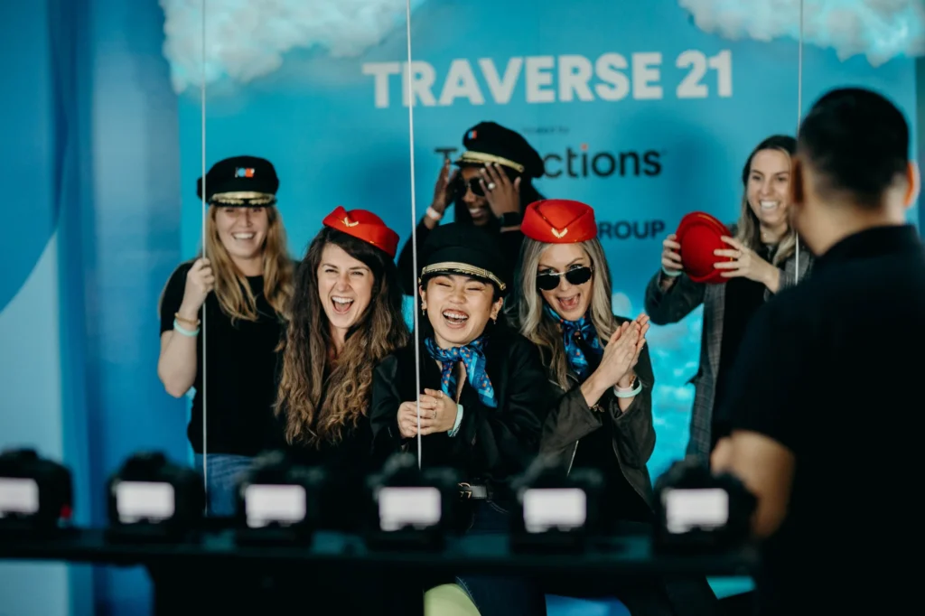 Six people pose cheerfully before a blue “TRAVERSE 21” backdrop, four wearing pilot or flight attendant hats. They laugh and smile as cameras capture the lively scene—perfect moments showcasing expert event planning in action.