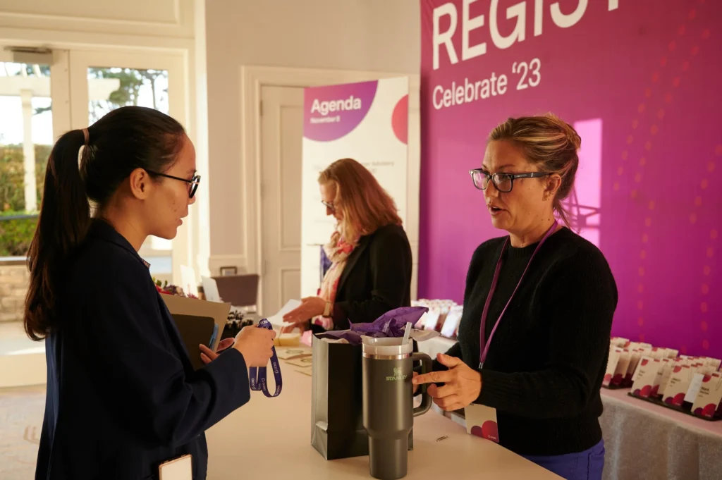Two women talk across a registration table at an event planned by a full-service event company. One hands over a lanyard as another arranges name badges beneath a purple “REGISTER” banner, showcasing expert event planning in action.