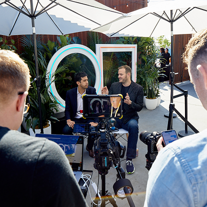 Two men sit on a white bench under umbrellas, being filmed and interviewed outdoors by a full-service event company. Cameras, a tablet, and crew members are visible as conference planning services bring the lush, neon-lit setup to life in the bright sunlight.