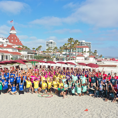 A large group of people in colorful shirts pose together on a sandy beach, raising their hands. Behind them are red umbrellas, palm trees, and historic hotel buildings—an ideal scene for any full-service event company specializing in memorable gatherings.