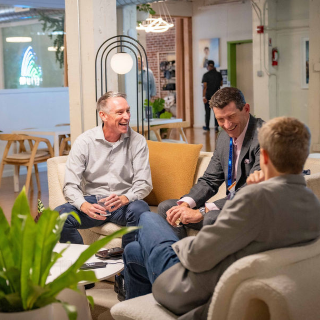 Three men in business attire sit and laugh together in a modern office lounge with cozy chairs, plants, and warm lighting—an ideal setting for conference planning services, fostering a friendly, collaborative atmosphere seen through large windows in the background.