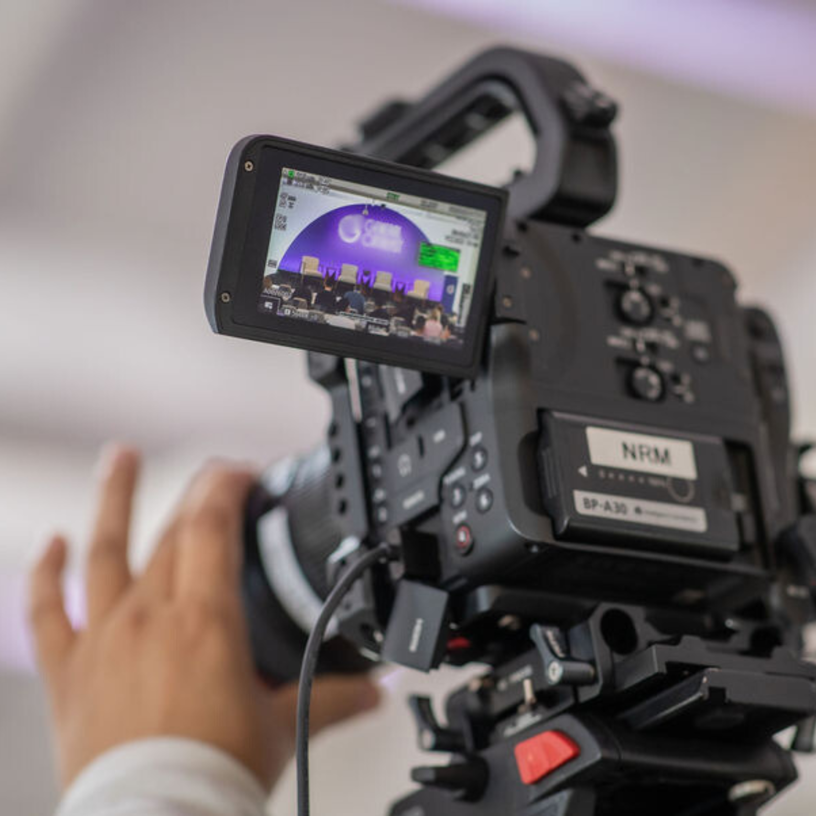 A close-up of a professional video camera being operated by a person. The camera’s display screen shows a live feed of a stage set for an event, highlighting the expertise of a full-service event company in conference planning services.