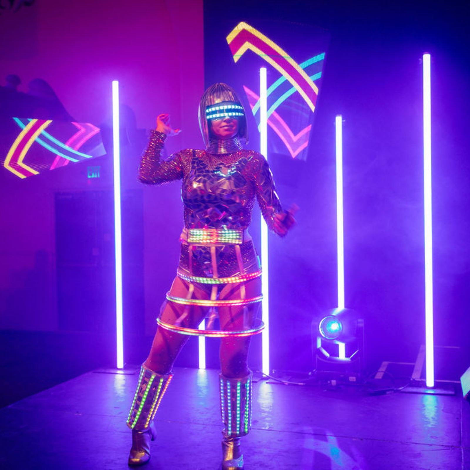 A performer in a futuristic outfit with glowing neon lights stands on stage—a dazzling highlight for any event planning. She wears a metallic dress, illuminated helmet, and LED boots as vertical purple light bars shine behind her in the smoky darkness.