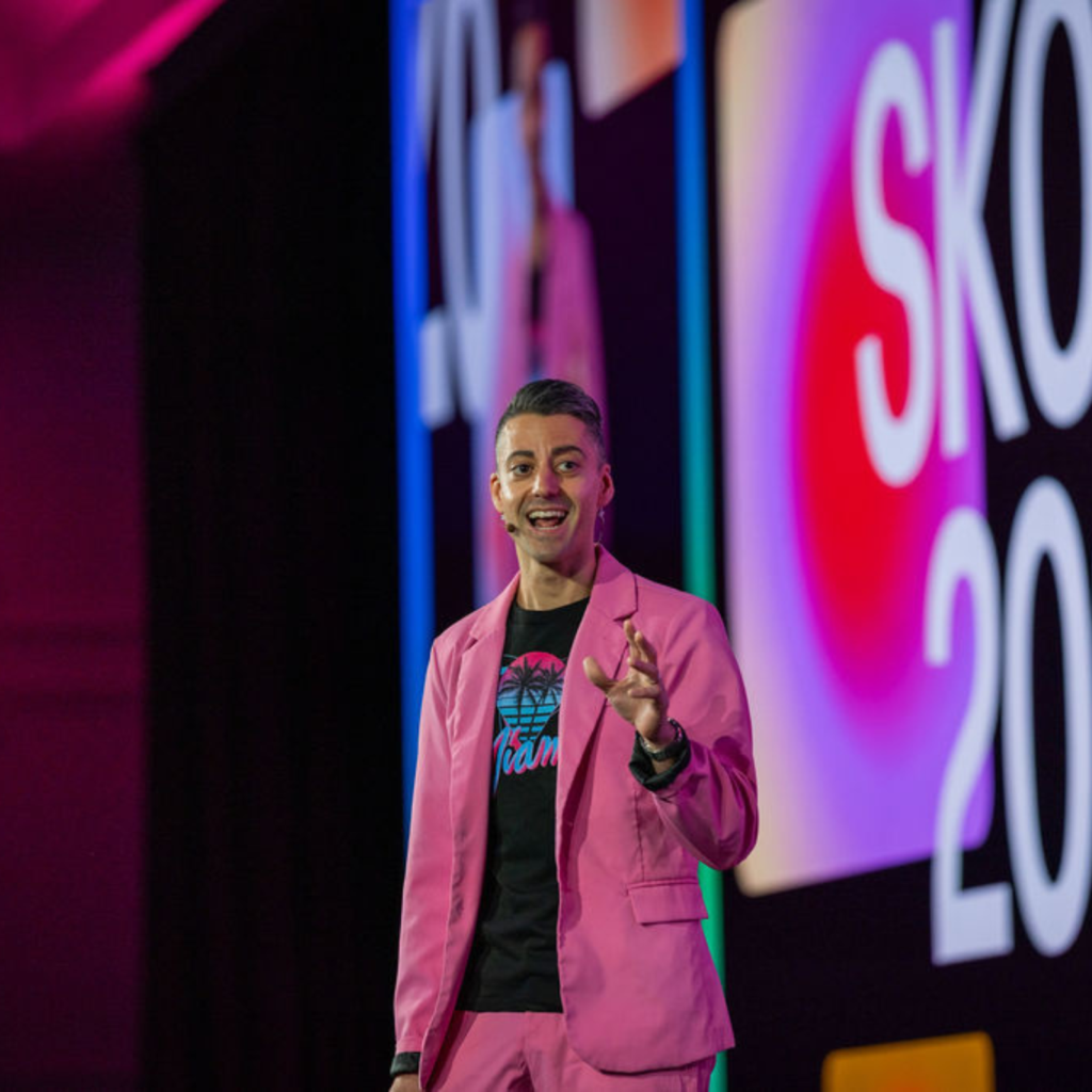 A person in a bright pink suit and black t-shirt is speaking on stage, gesturing with their right hand. Behind them, a bold SKO 2023 display pops as vibrant lighting sets the scene—perfect inspiration for virtual event planning.