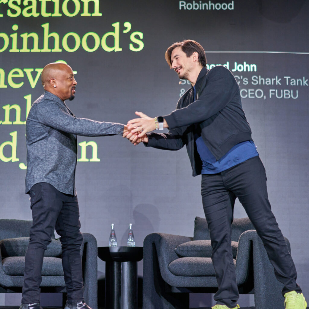 Two men stand on stage shaking hands and smiling, exemplifying successful event planning. One wears a dark zip-up jacket and sneakers, the other a gray shirt and black pants. Behind them, bold yellow and white text is visible above empty chairs and water bottles.