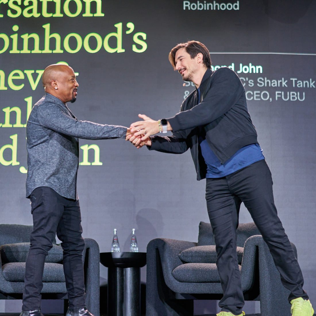 Two men stand on stage shaking hands and smiling, exemplifying successful event planning. One wears a dark zip-up jacket and sneakers, the other a gray shirt and black pants. Behind them, bold yellow and white text is visible above empty chairs and water bottles.