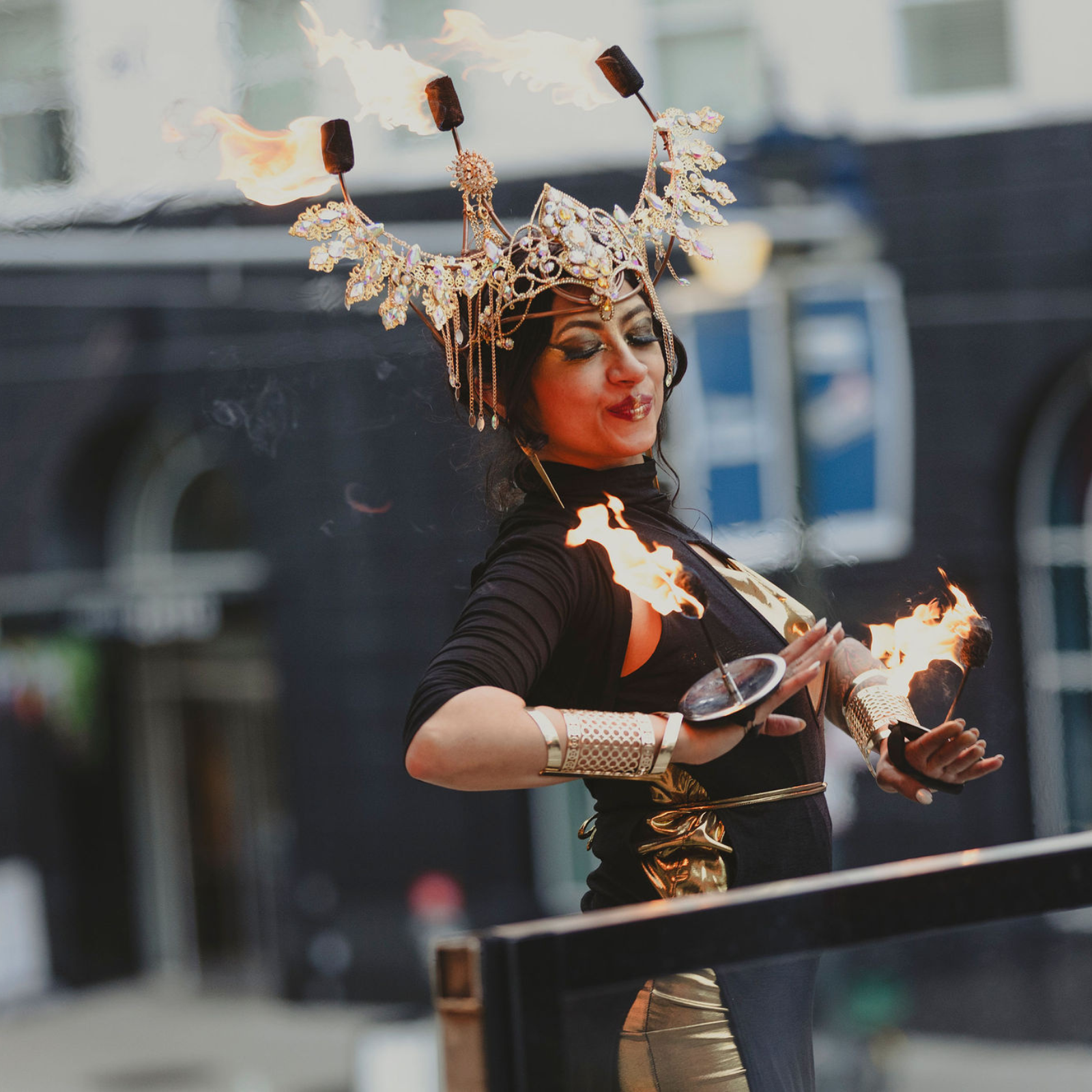 A woman in gold and black costume performs a fire dance outdoors—perfect inspiration for virtual event planning. Her jeweled headpiece, flaming wands, and dramatic makeup create a festive, mystical look against blurred buildings.