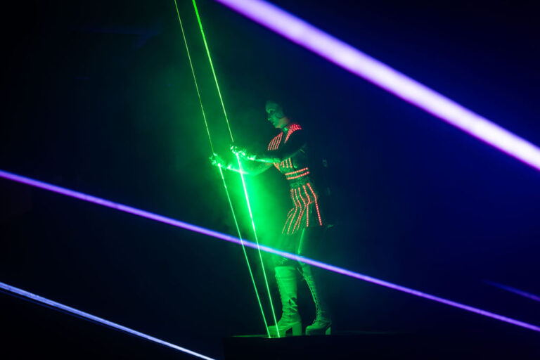 A person in illuminated, futuristic clothing interacts with bright green laser beams on a dark stage. As purple and blue lasers cross the scene, fog enhances the vibrant sci-fi atmosphere—perfect inspiration for innovative event planning.