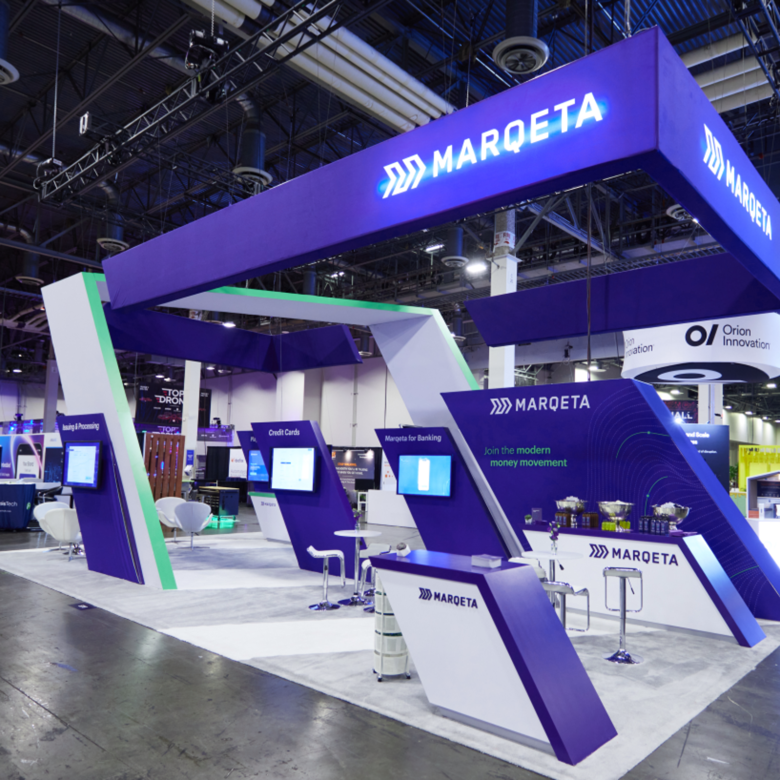 A modern, spacious trade show booth for Marqeta features bold purple, blue, and white branding, angled structures, digital screens, stools, and counters. Overhead signs display the Marqeta logo. A full-service event company can elevate such conference experiences.