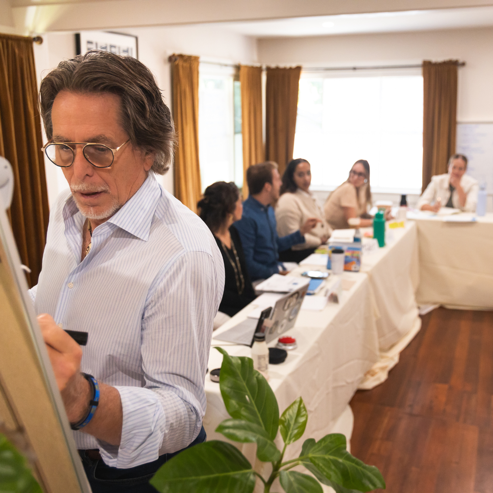 A man with glasses writes on a whiteboard in a bright room. Five people sit at a table with laptops, notebooks, and drinks, watching him as they discuss event planning. The space features wooden floors, green plants, and golden curtains lit by sunlight.