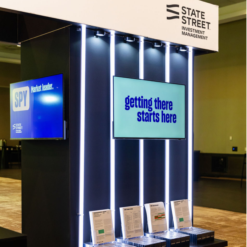 A modern exhibition booth for State Street Investment Management features vertical LED lights, a large screen displaying “getting there starts here,” and brochures on shelves below—ideal for event planning and conference planning services.